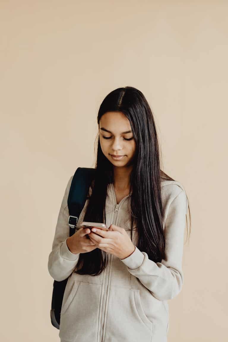 Teenage girl with long hair using a smartphone, wearing a backpack, against a beige background.